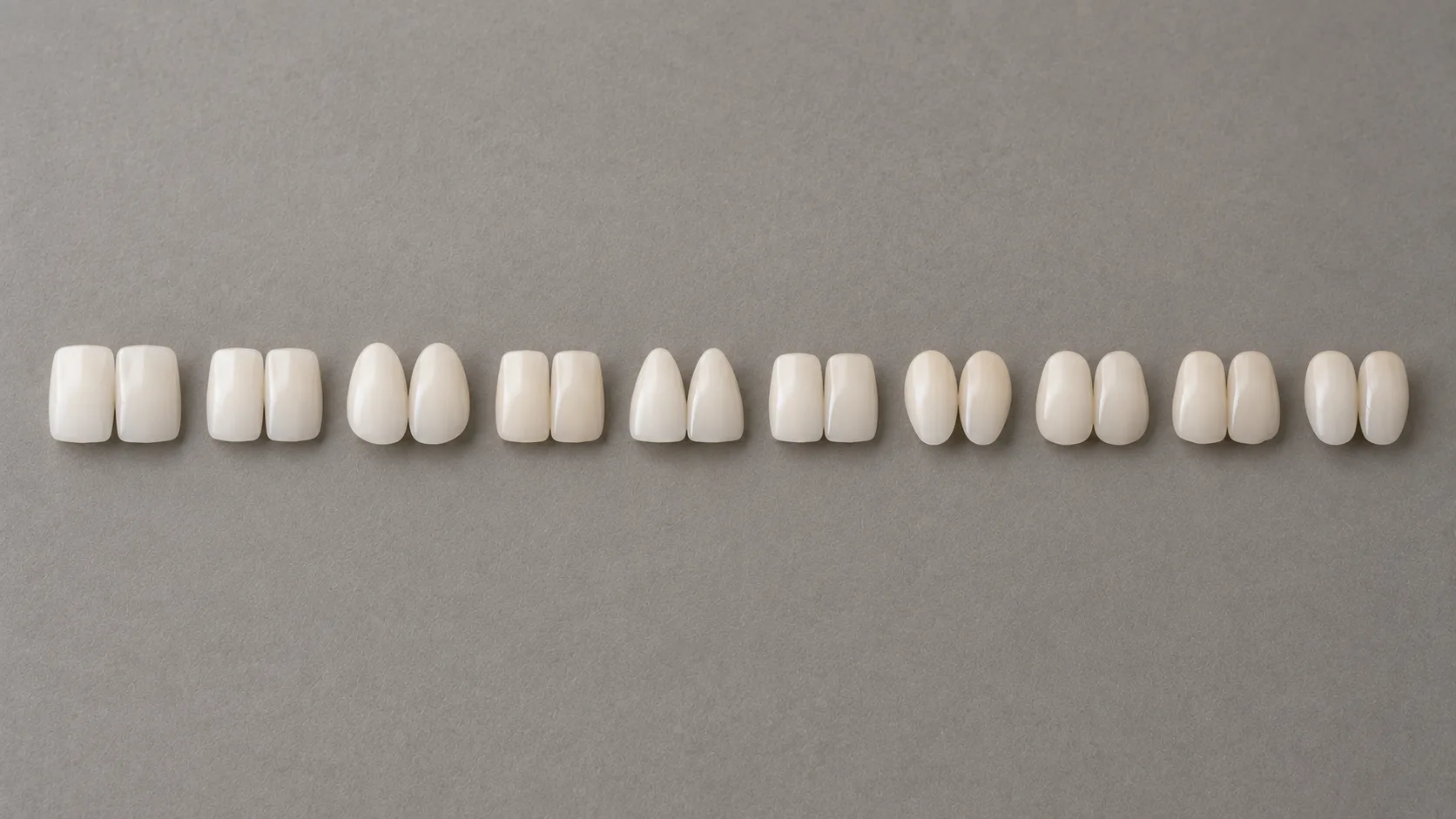 A long row of porcelain front teeth of different shapes laid evenly across grey card
